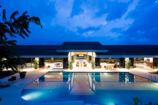 The image shows a luxurious building with a large, illuminated swimming pool in the foreground against a twilight sky.