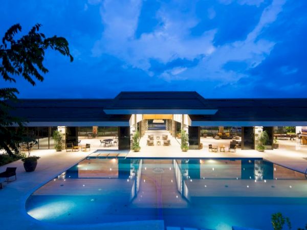 The image shows a luxurious building with a large, illuminated swimming pool in the foreground against a twilight sky.
