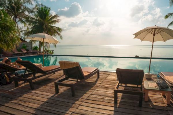 A serene beachside pool area features lounge chairs, umbrellas, and a stunning ocean view under a partly cloudy sky.