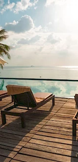 A serene coastal scene with lounge chairs and umbrellas on a wooden deck overlooking an infinity pool, and the ocean beyond under a sunny sky.