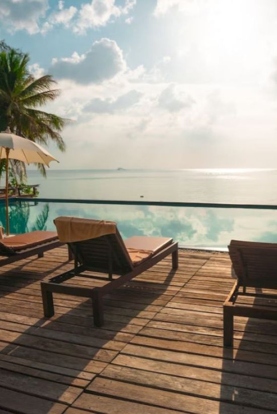 A serene poolside setting with lounge chairs, umbrellas, and palm trees overlooks a calm sea under a partly cloudy sky.