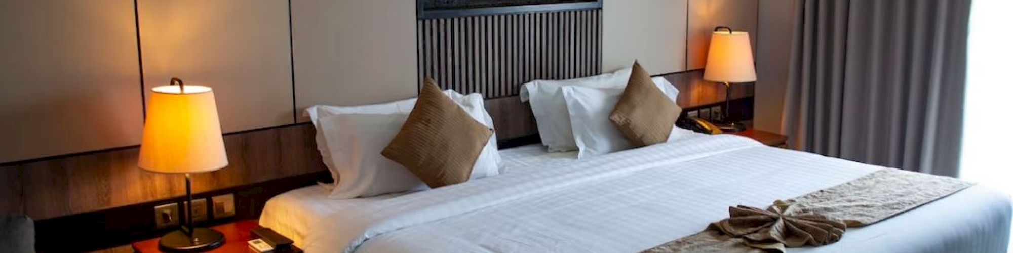 The image shows a modern hotel room with a large bed, two bedside tables with lamps, decorative pillows, and a wall-mounted picture above the bed.