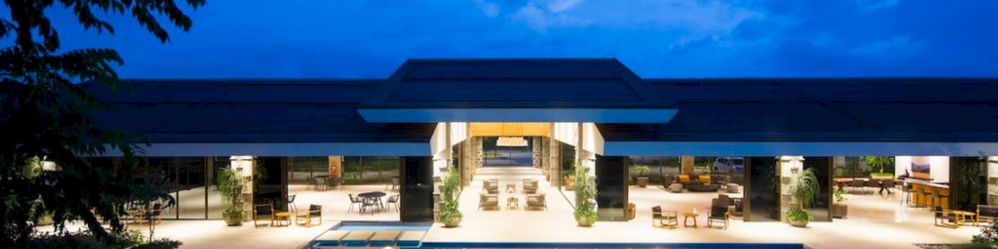 A luxurious modern building featuring a large, illuminated swimming pool in front, surrounded by outdoor seating and vegetation, under a twilight sky.