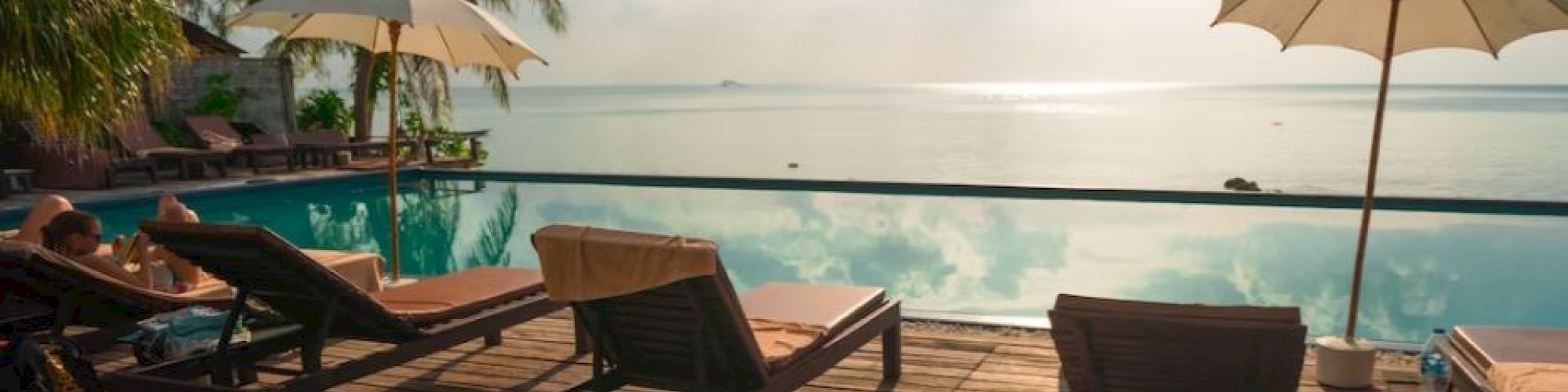 A scenic poolside view with lounge chairs, umbrellas, and palm trees overlooking a calm sea under a partly cloudy sky, suggesting a tropical paradise.