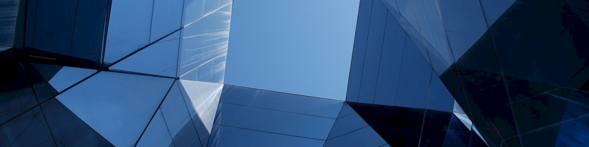 The image shows an upward view of a geometric, modern glass building with reflective surfaces, creating a kaleidoscopic effect against the sky.