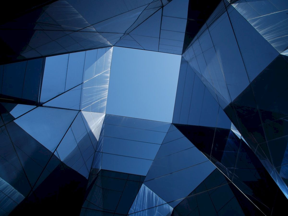 The image shows an abstract view of modern architecture with glass panels reflecting blue sky, creating geometric patterns.