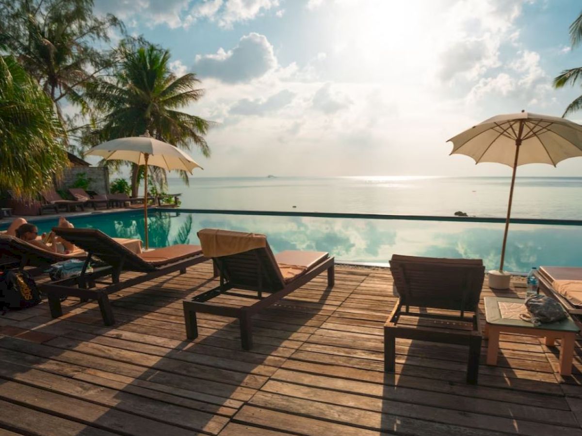A serene poolside scene with lounge chairs, umbrellas, palm trees, and a stunning view of the water under a bright, cloudy sky is visible.