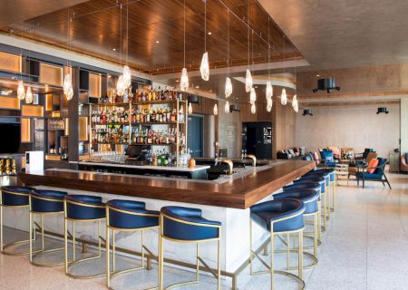 A modern bar with a wooden counter, blue chairs, hanging lights, and a stocked shelf of bottles. There is seating and a lounge area in the background.