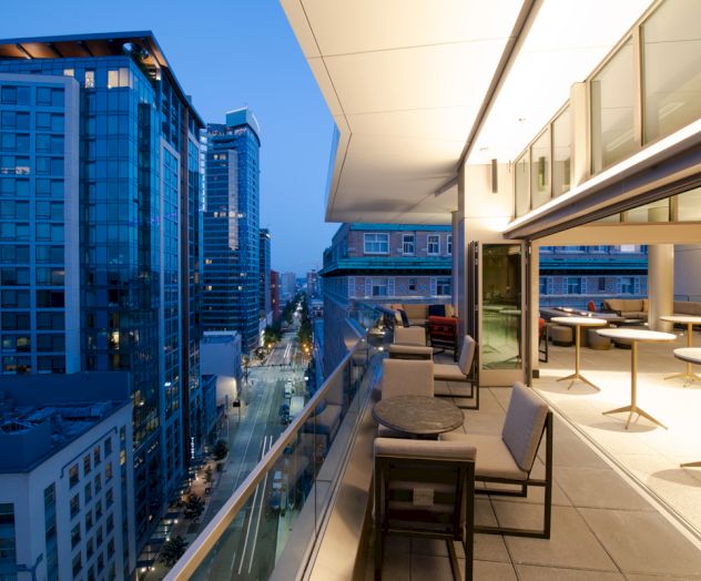 A modern cityscape from a high-rise balcony, featuring tall buildings, a street below, and elegant outdoor seating on the terrace.