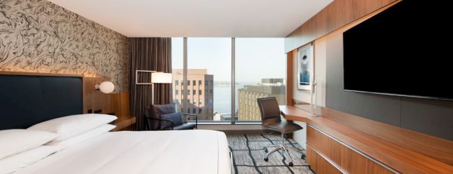 A modern hotel room features a large bed, wall-mounted TV, desk with a chair, stylish wallpaper, and a window with a city view.