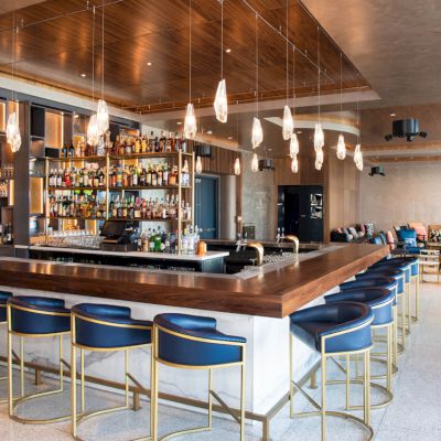 This image shows a modern bar with a wooden countertop, blue bar stools, hanging lights, and a well-stocked liquor shelf, adjacent to a lounge area.