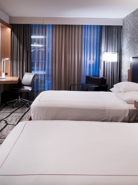 A modern hotel room with two beds, a wall-mounted TV, a desk by a window, and stylish lighting and decor.