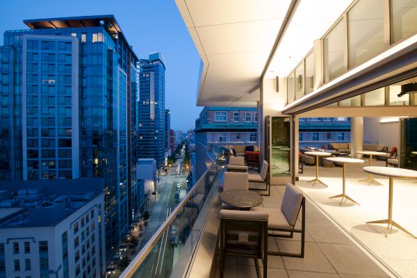 A modern balcony with chairs and tables, overlooking a city street lined with tall buildings during dusk, with some lights on.