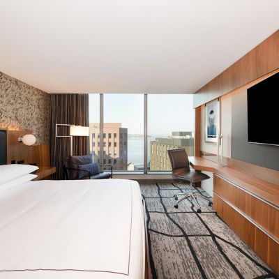 A modern hotel room features a large bed, desk, chair, wall-mounted TV, and floor-to-ceiling windows with a city view.
