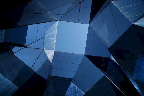 The image shows a view looking up at modern glass buildings with reflective surfaces forming geometric shapes against the blue sky.