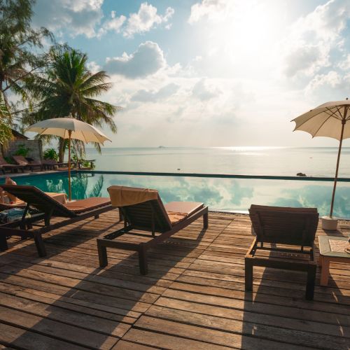 The image shows a serene poolside scene with lounge chairs, umbrellas, and a beautiful ocean view under a partly cloudy sky with sun shining.