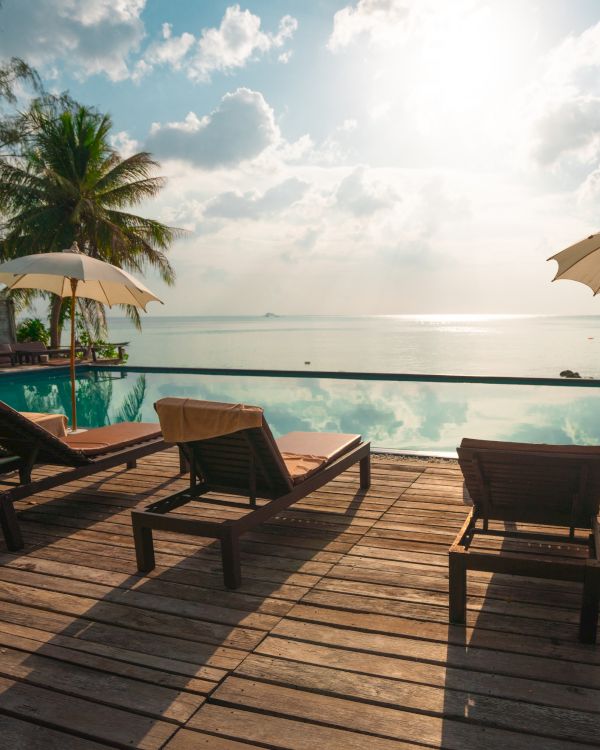 The image shows a serene poolside scene with lounge chairs, umbrellas, and a beautiful ocean view under a partly cloudy sky with sun shining.