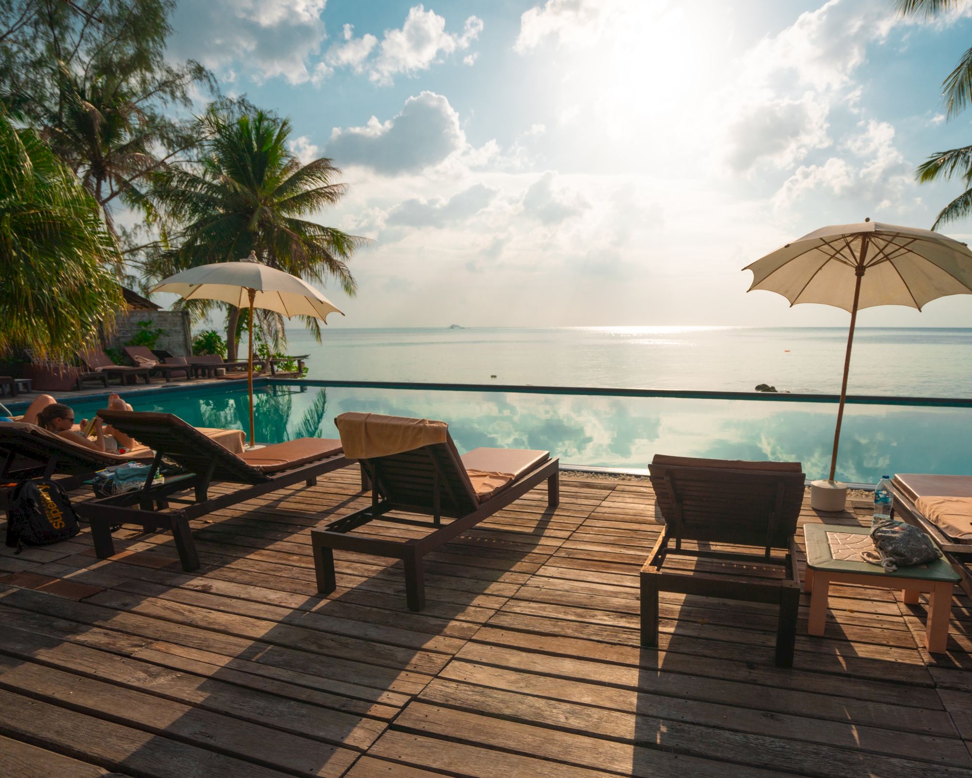 The image shows a serene beachside scene with lounge chairs, umbrellas, a wooden deck, and palm trees, overlooking a calm ocean under a bright sky.