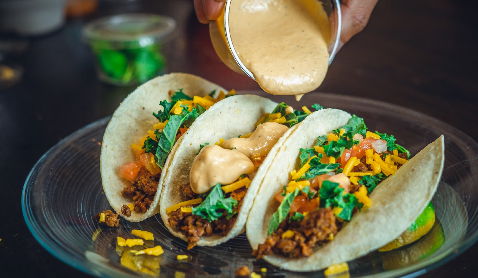 The image shows three tacos filled with seasoned meat, lettuce, tomatoes, cheese, and a sauce being poured on top, all on a clear plate.