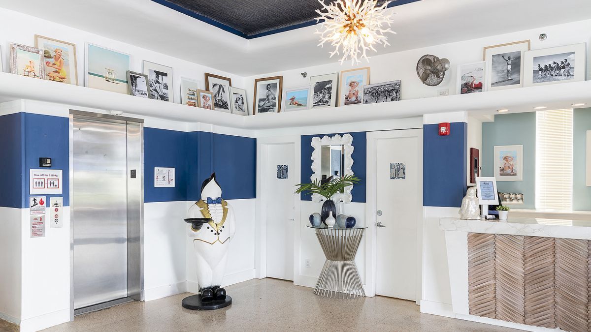 The image shows a stylish lobby with a blend of blue and white decor, featuring an elevator, framed pictures, a sculpted penguin, and a reception desk.