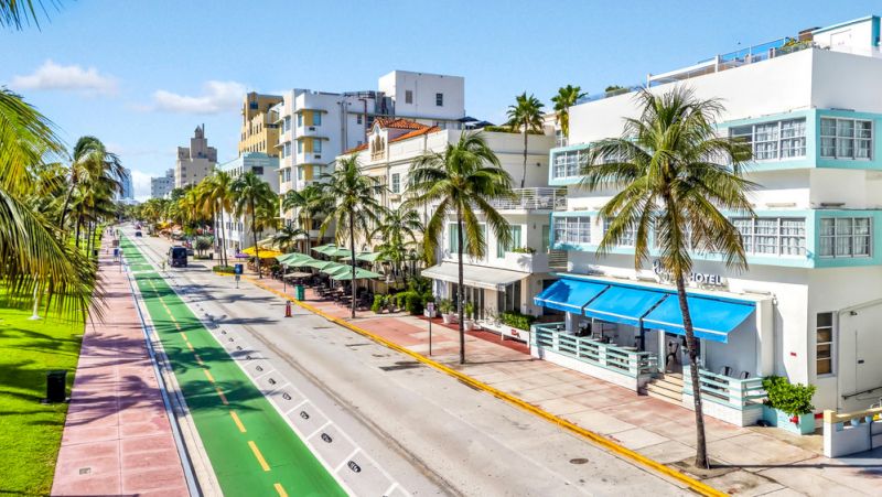 A white hotel building with blue accents and palm trees in front of it on a street with a bike lane.