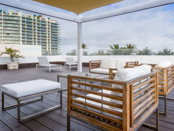 An outdoor patio with modern wooden furniture, white cushions, lounge chairs, and potted plants overlooking nearby buildings and palm trees.
