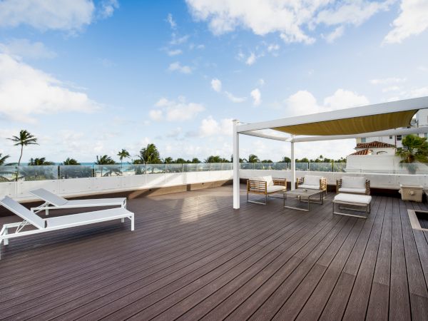 A spacious outdoor deck with lounge chairs, seating arrangements under a pergola, and a scenic view of palm trees and a blue sky with clouds.