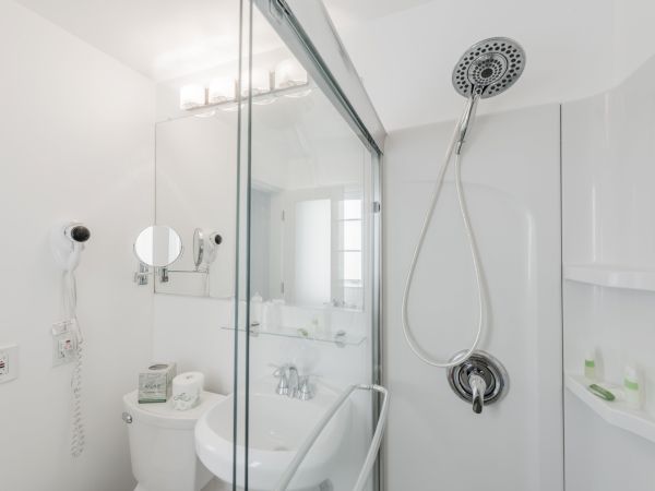 A modern bathroom with a shower, sink, mirror, and toilet. It has white walls and fixtures, a wall-mounted hair dryer, and shelf for toiletries.