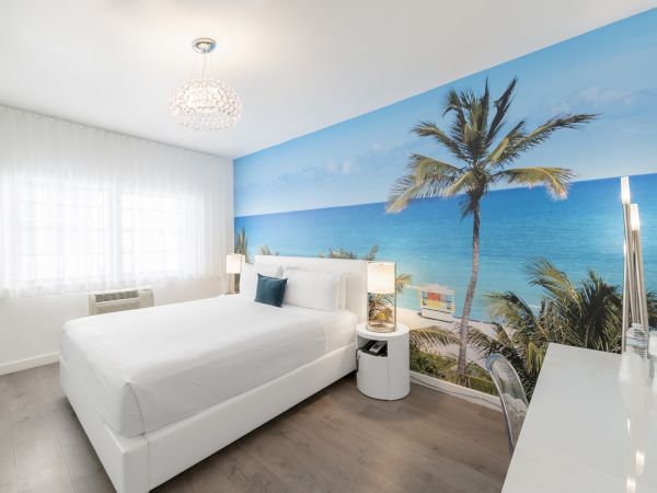 A modern bedroom with a white bed, a chandelier, and a wall mural depicting a tropical beach scene with palm trees and clear blue water ending the sentence.