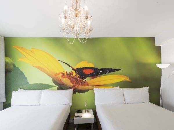 The image shows a hotel room with two beds, a chandelier, a floor lamp, and a large mural of a butterfly on a yellow flower behind the beds.
