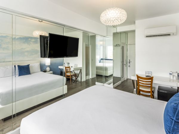 A modern hotel room featuring two beds with blue pillows, a large TV, mirrored walls, a desk with a chair, and an air conditioning unit.