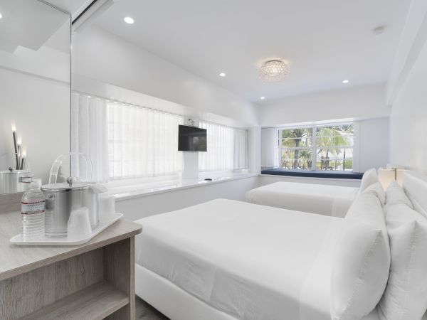 A modern hotel room features two beds, a flat-screen TV, a window view, and a small desk with water bottles and cups.