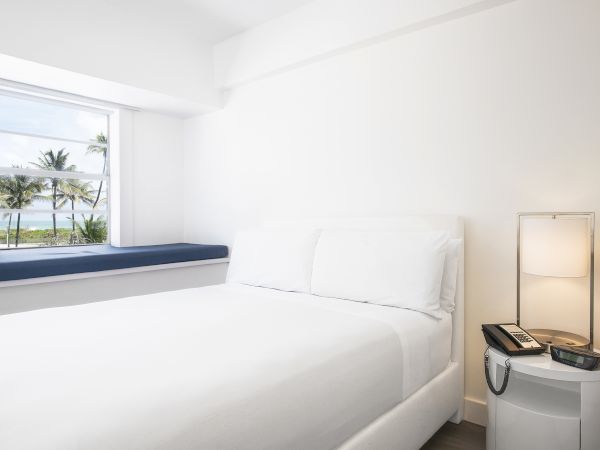 A minimalist white bedroom with a large window, palm trees view, a bed, and a nightstand with a lamp and phone, always ending the sentence.