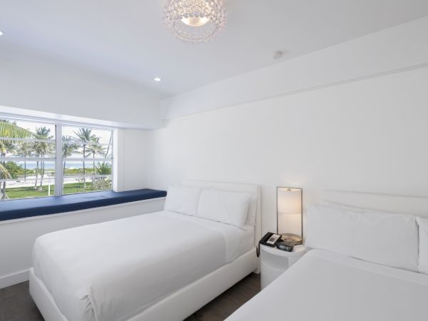 A bright, clean hotel room with two white beds, a window seat, a bedside table with a lamp, and a large window overlooking palm trees and water.