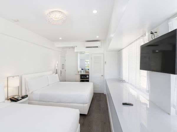 A bright, modern hotel room features two beds, a wall-mounted TV, nightstands with lamps, and a kitchenette area.