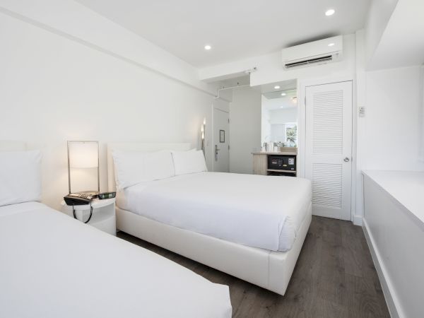 A modern, white-themed hotel room with two beds, a nightstand with a lamp, an air conditioner, a small kitchen area, and minimal decor.