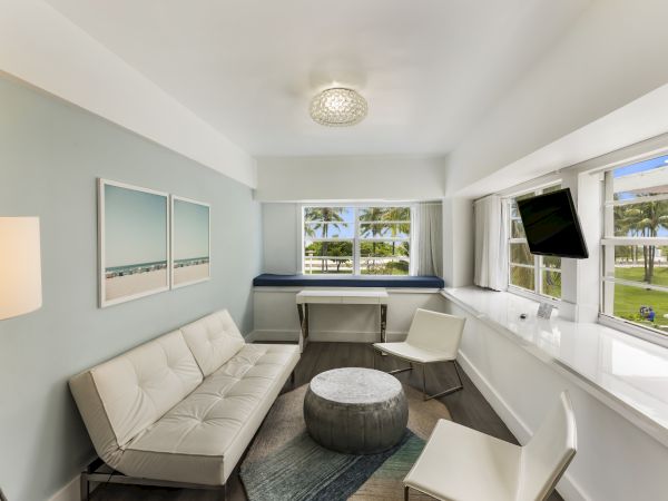 The image shows a modern living space with a sofa, two chairs, a small table, and a wall-mounted TV. Large windows provide a view of palm trees outside.