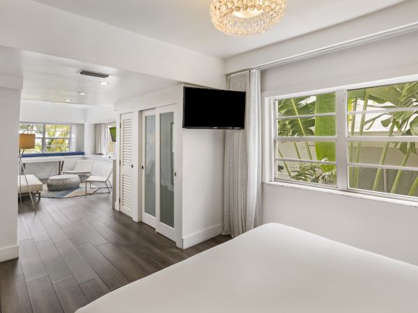A modern, bright bedroom with a large window, flat-screen TV, and adjacent seating area, featuring stylish, minimalistic décor and wooden flooring.