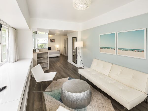 A modern living room with a white sofa, chair, round silver ottoman, and TV, featuring beach-themed artwork on the wall and a lamp in the corner.