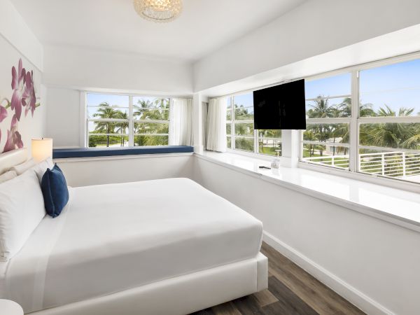 A modern bedroom with a white bed, a floral wall design, large windows with a view, a TV, and a blue bench by the window at the far end.