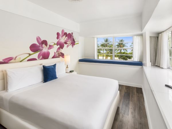 A bright, modern bedroom with a large bed, floral wall art, window seat, and a scenic view of palm trees through the window, with a TV on the wall.