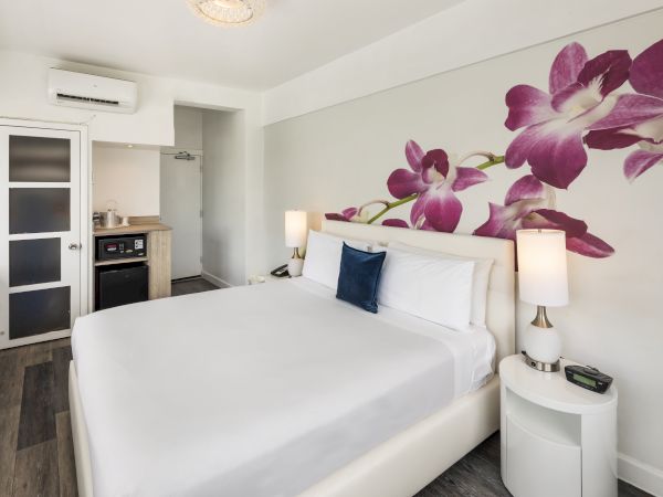 A modern hotel room with a bed, purple floral wall art, two bedside lamps, an air conditioner, a small kitchenette, and wooden flooring.