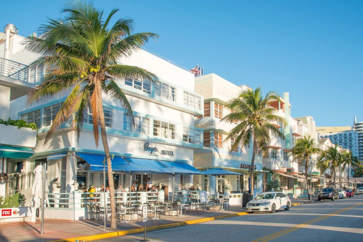 A white, multi-story building with blue accents and awnings, featuring outdoor seating and palm trees on a sunny street.
