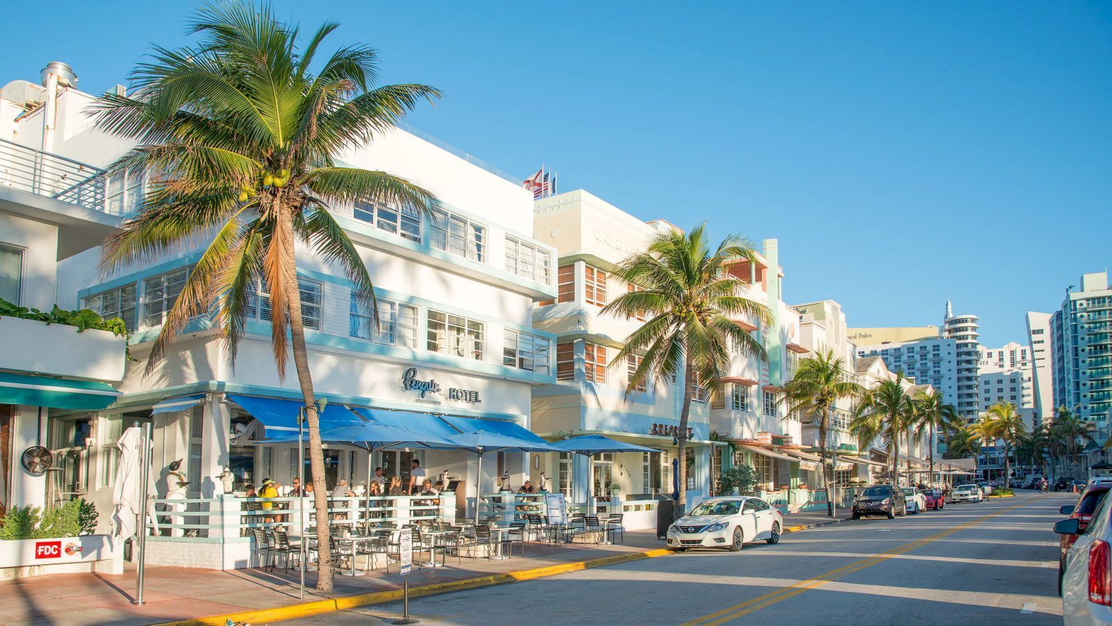 A sunny street with palm trees, art deco buildings, outdoor cafes, cars parked along the road, and people enjoying a leisurely day.