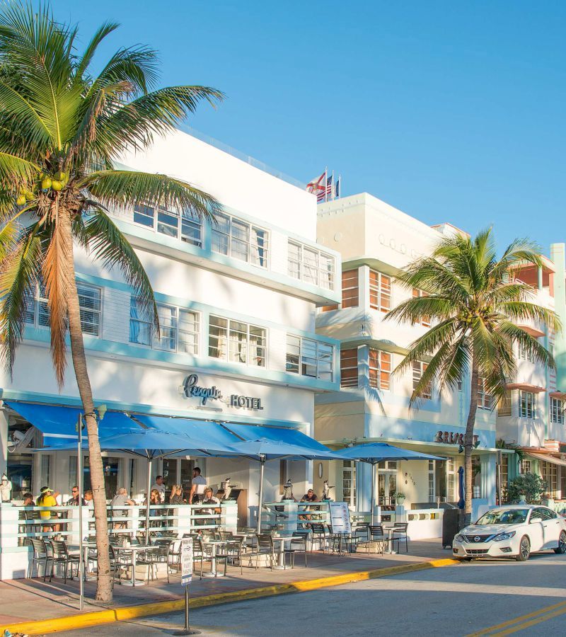 A white, three-story building with blue awnings and palm trees in front of it.