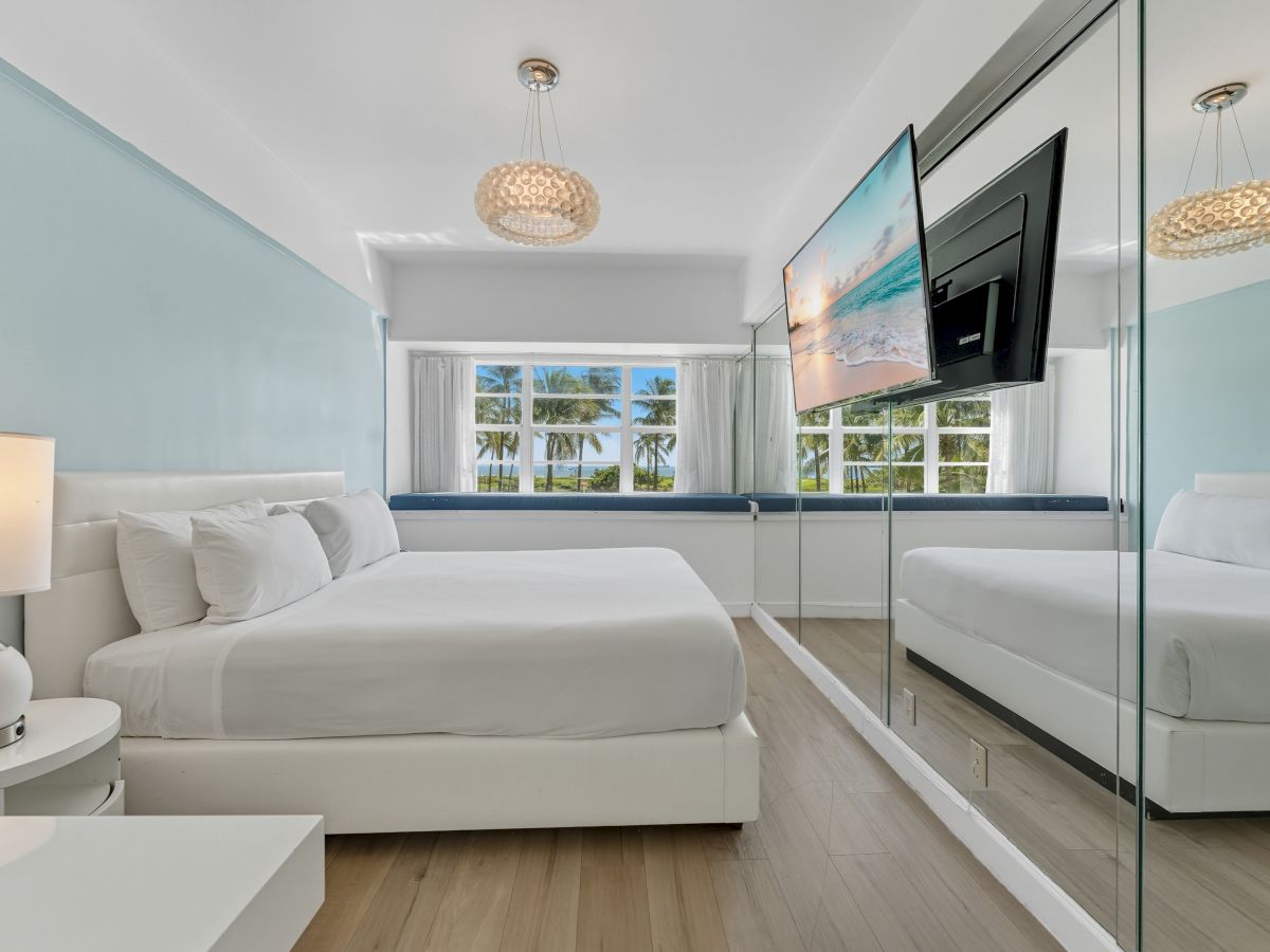 A modern bedroom features a large bed, mirrored wall, TV, and elegant lighting, creating a clean and stylish atmosphere.