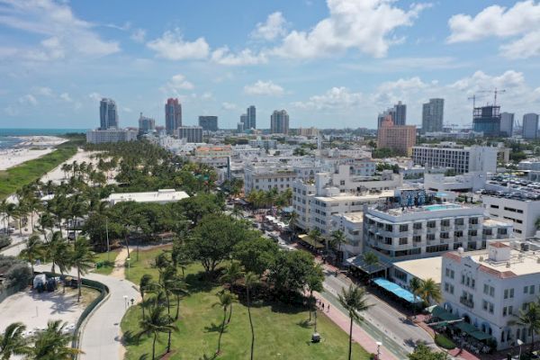 The image shows an aerial view of a coastal city with numerous buildings, palm trees, a park, and a beach in the background.