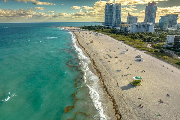 An aerial view of a beach with turquoise water, a sandy shore, beachgoers, and tall buildings in the background under a partly cloudy sky.