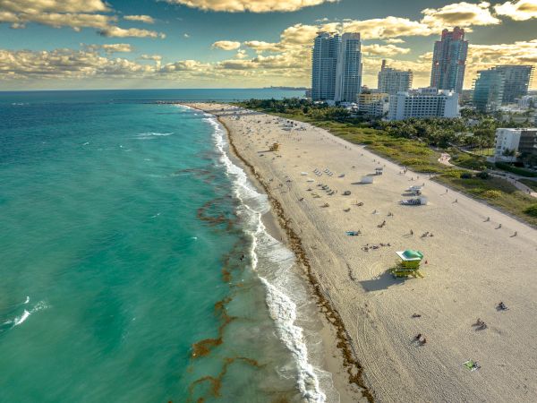 An aerial view of a beach with turquoise water, a sandy shore, beachgoers, and tall buildings in the background under a partly cloudy sky.