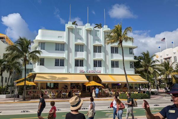 A street scene with people walking and a large Art Deco-style building, palm trees, and a restaurant with a yellow canopy are visible.